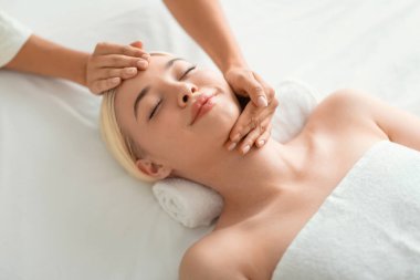 Beauty And Relaxation. Young blonde woman enjoying facial massage, lying on spa bed indoor at beauty salon, high angle shot of lady relaxing during facebuilding treatment