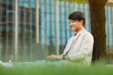 Asian guy freelancer sitting on green lawn under tree at public park, working on laptop. Side view chinese young man student studying online at university campus, typing on computer keyboard