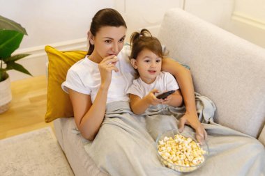 Mom and daughter watching TV with popcorn on sofa, relaxed young mother and preschooler female child lying under blanket, bonding together while resting in living room at home,