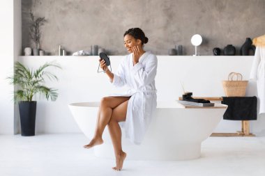 Attractive young black woman in white bathrobe, sitting on the edge of bathtub and looking into handheld mirror in chic bathroom, happy african american female enjoying her beauty after skincare