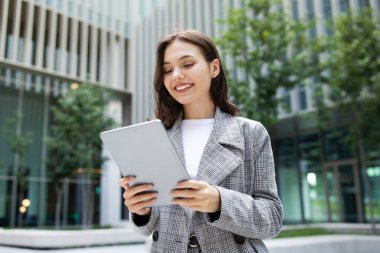 Smiling business lady happily browsing on digital tablet computer, standing outside an urban office building, showcasing successful work lifestyle with modern online technology