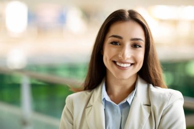 Close-up of a radiant happy caucasian young businesswoman with a friendly smile, wearing a light beige suit jacket and glasses, in a modern corporate office setting, close up