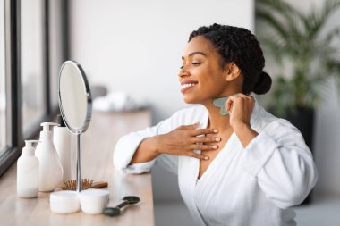 Beautiful young black woman doing neck massage with gua sha tool, smiling attractive african american lady massaging skin while sitting near mirror at home, making beauty treatments after bath
