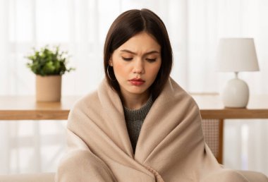 Closeup of upset beautiful brunette young woman wrapped in blanket sitting on couch, freezing at home, feeling cold during winter season in unheated apartment
