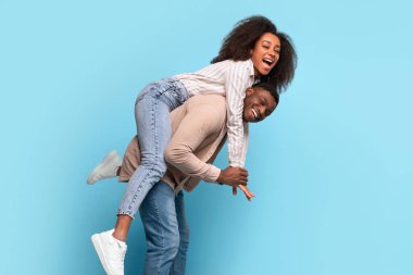 Laughing black woman enjoying piggyback ride from smiling man, both having great time with playful gestures against a light blue backdrop
