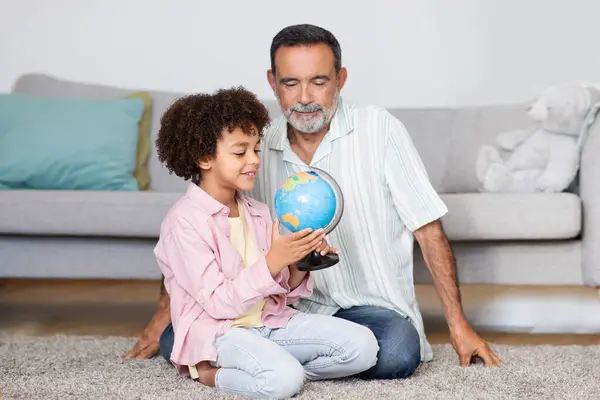 Mature Grandpa And His Little Grandson Holding World Globe, Learning Geography Lesson And Talking About Traveling, Sitting On Floor At Home Interior, Bonding With Grandchild On Weekend