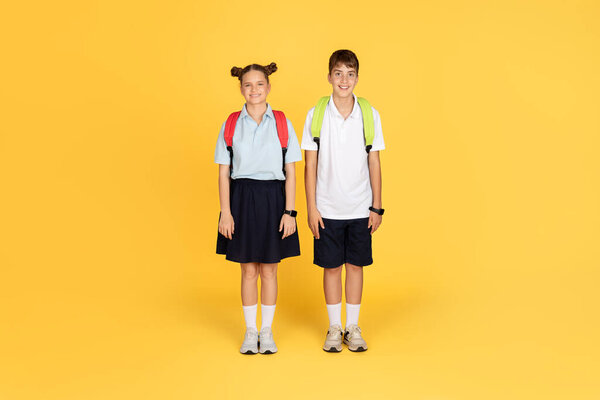 Two cheerful schoolchildren, a boy and a girl, wearing uniforms and backpacks, standing confidently with bright smiles on a sunny yellow background, ready for a new school day