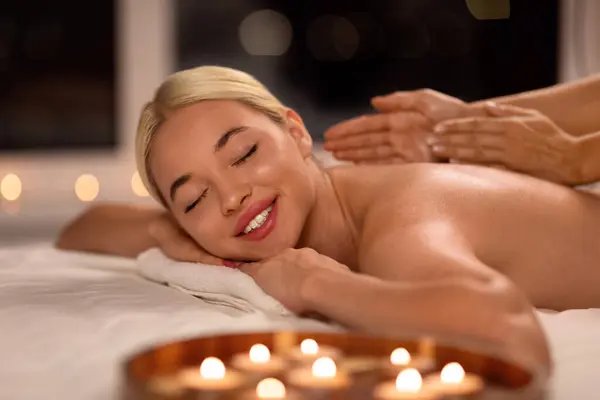 Young Blonde Lady During Massage Therapy Relaxing And Enjoying Procedure, Receiving Professional Back Massage From Masseur, Lying On Bed In Spa Salon With Burning Candles, Eyes Closed