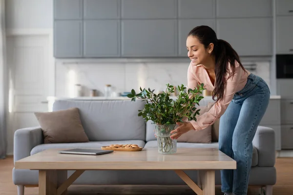 Smiling woman in peach blouse and blue jeans carefully arranges plant in vase on wooden coffee table in bright, modern living room setting