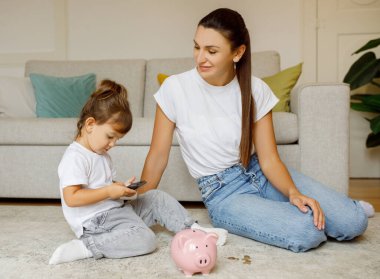 Children And Money Management. Little girl counting coins from pink piggybank and using calculator, cute preschool female child sitting next to mom in living room, mother teaching daughter savings