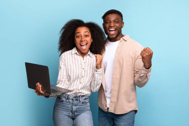 Young and energetic black couple holding open laptop, jubilantly celebrating win or good news, against vibrant blue background