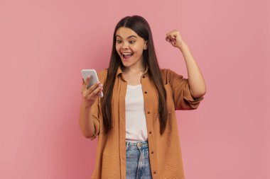 Joyful teenage girl looking at her smartphone and shaking fist, happy teen female expressing excitement or success, posing against pink studio background, embodying positive digital interaction
