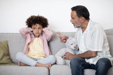 Senior Grandpa Scolding Naughty Grandchild Boy For Misbehavior While Grandson Covering His Ears, Sitting On Sofa In Living Room At Home. Bad Multigenerational Relationships, Family Problems