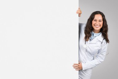 Medical ad. Happy european woman doctor peeking out of huge blank advertisement board, therapist in uniform demonstrating empty space on white placard