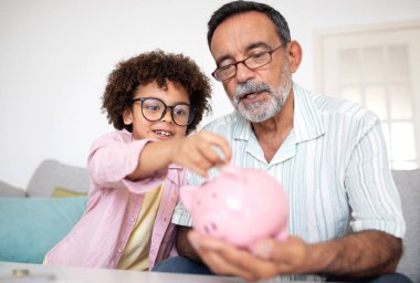 Family Finance Lessons. Hispanic Grandfather And Grandchild Boy Sharing Joy of Saving, Putting Money In Piggybank, Sitting On Sofa In Modern Living Room Indoors, Investing in Kids Financial Future