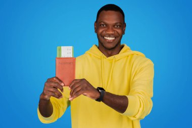 A smiling Black man in a yellow hoodie happily displays his passport and boarding pass, signifying the joys of international travel and adventure on a bright blue background