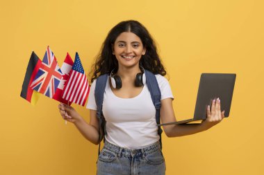 Happy young indian woman student with many different flags holding computer, isolated on yellow background, studio. Online school, course, education, ad, offer, modern technology