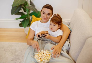 Mom and cute little daughter watching TV with popcorn on sofa, smiling family mother and preschooler female kid relaxing together at home, resting on comfy couch, child holding remote controller