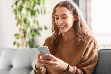 Young teen lady texting friends using cellphone sitting on couch in her room at home, comfortably engaged with her mobile phone, enjoying games and social media apps indoors