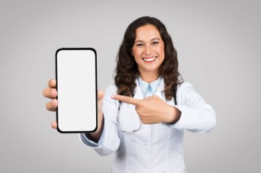 Cheerful female doctor in lab coat showing a mobile phone with a blank screen, pointing at it with her finger, on neutral grey background, mockup