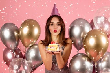 A surprised woman with a party hat is playfully blowing out a candle on a slice of cake, surrounded by a festive ambiance of metallic balloons and confetti on a pink backdrop