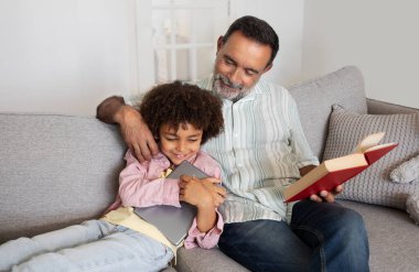 Hispanic grandfather and grandchild spending time with digital tablet computer and paper book together, happily scrolling and reading on weekend, sitting together on couch at home