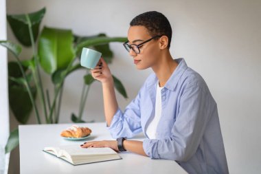 A contemplative latin young woman enjoys her morning coffee while reading, surrounded by the lush greenery of indoor plants, creating a serene atmosphere. Work, study, relax