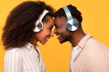 Intimate and joyful moment as young black couple in wireless headphones, touching foreheads and smiling, standing on yellow background, side view