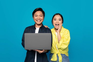 Surprised and happy millennial Asian couple with a laptop, the woman touching her face in excitement and the man smiling, indicating good news or success on a cyan background