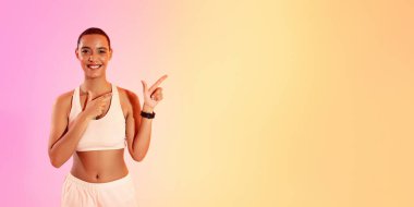 An engaging woman with a shaved head points to her left with a bright smile, dressed in a white sports bra and pale pink shorts against a soft gradient background, panorama