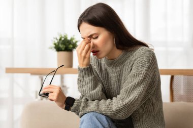 Sleepy young caucasian woman holding glasses rubbing eyes, feels tired after working on computer, sitting on couch at home. Exhausted lady suffering from ocular diseases, eye strain.