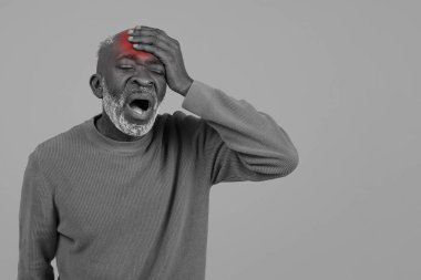 Black and white image of a senior Black man with a vivid red spot on his head, holding his head in pain or distress, wearing a sweater, isolated on a grey background