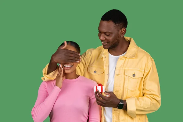 Black guy make anniversary surprise to his girlfriend, young man close woman eyes, holding little gift box with jewelry engagement ring, green background. Proposal, Valentine Day celebration