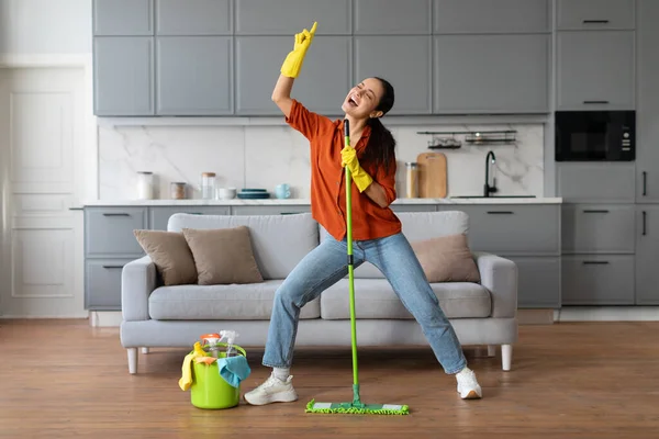 Playful woman in orange shirt and denim jeans, with yellow gloves, dances with green mop by sofa and bucket of cleaning tools, enjoying housework