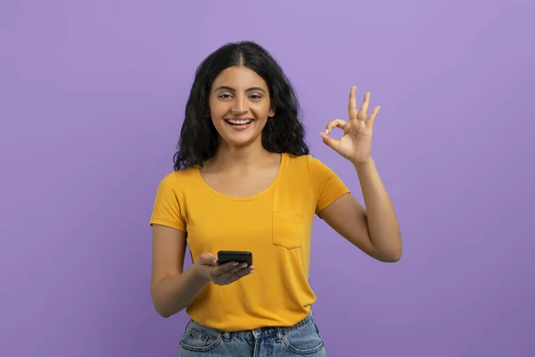 Nice educational mobile application, online studying. Cheerful attractive curly young hindu lady with smartphone in her hand showing okay gesture and smiling, purple studio background
