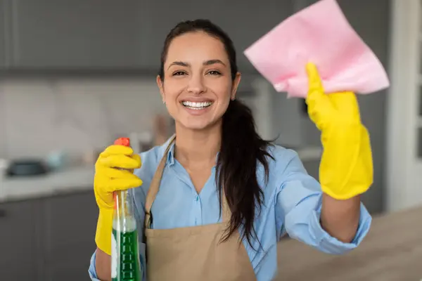Smiling young woman with yellow gloves holding green cleaning spray bottle and pink cloth, ready to clean, with beige apron in modern kitchen