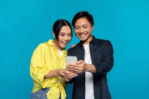 Enthusiastic Asian couple sharing a joyous moment while jointly looking at a smartphone screen, possibly enjoying an entertaining app or an amusing message, against a vivid cyan background