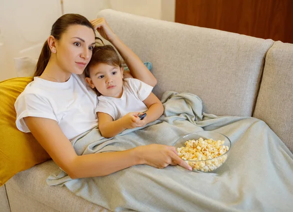 Family Of Two, Single Mother And Little Daughter Relaxing On Couch At Home Together, Young Mom And Cute Preschooler Female Child Lying On Sofa Under Blanket, Eating Popcorn And Watching Tv