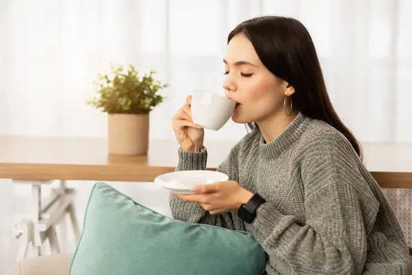Relaxed beautiful brunette young hispanic woman wearing warm sweater enjoying aromatic coffee at home, sitting on couch in cozy living room, sipping hot tea, enjoying delicious drink, copy space