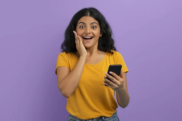 stock image Discount, promotion, online offer, sale. Emotional excited young indian woman with phone in her hand touching her face and gesturing, isolated on purple background