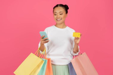 Smiling Asian Lady Holding Smartphone, Credit Card And Shopping Bags, Happy Korean Woman Making Online Shopping In Mobile App While Standing Isolated Over Pink Studio Background, Copy Space