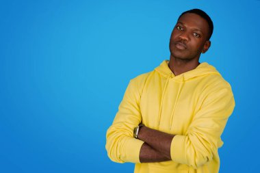 A confident calm african american man student stands with arms crossed wearing a yellow hoodie, looking thoughtfully at the camera against a uniform blue studio background