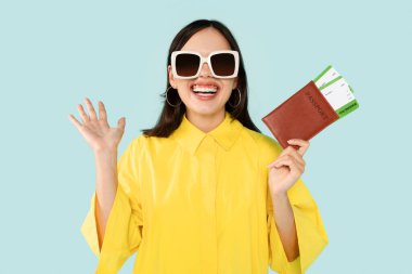 Super happy and excited young woman going vacation, showing passport and flight tickets boarding pass, wearing yellow shirt and sunglasses, gesturing and smiling, blue studio background