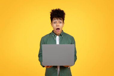 Online education, e-learning. Amazed young black guy using laptop, typing on computer keyboard, looking at camera and grimacing, copy space for advertisement, yellow studio background