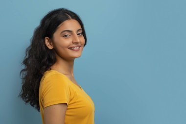 Profile portrait of attractive curly young indian woman wearing yellow t-shirt posing on blue studio background, smiling at camera, copy blank space for advertisement