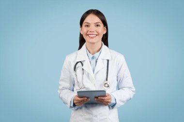 Cheerful female healthcare professional in a lab coat with a stethoscope around her neck holding a digital tablet against a blue background