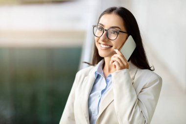 Cheerful caucasian young businesswoman, boss engaged in a phone conversation, wearing stylish glasses and a formal beige suit, in a well-lit office environmen, close up