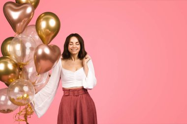 Contented caucasian glad young woman in a stylish white top and maroon skirt, holding a bunch of gold and clear balloons with a gentle smile, against a soft pink backdrop