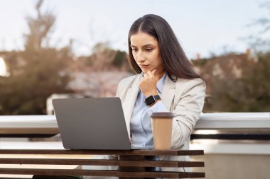 Focused businesswoman analyzing data on her laptop at a wooden outdoor table with a takeaway coffee cup, embodying professionalism and dedication in a relaxed environment