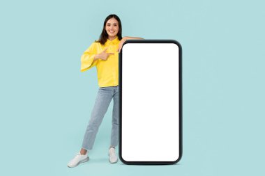 Cool cheerful stylish pretty young brunette woman pointing at huge phone with white blank mockup screen, showing nice online offer or recommending mobile app, blue background, copy space